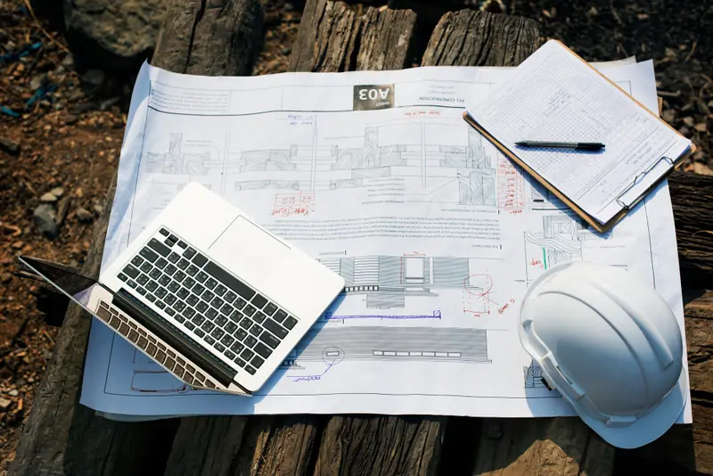 Planta de obra aberta com notebook, prancheta e capacete sobre madeira